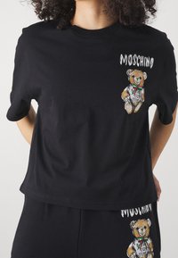 Černé bavlněné tričko s grafikou medvěda v kresleném stylu a textem "Moschino" na přední straně. Krátké rukávy, kulatý výstřih.
