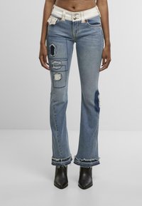 Jeans en denim bleu clair évasés avec des patchs déchirés et des ourlets effilochés. Possède une ceinture crème avec des détails de boutons et de poches.