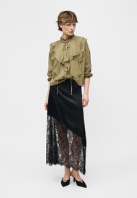 Vrouw met kort haar, gekleed in een olijfgroene blouse met ruches, een asymmetrische zwarte rok met een doorschijnende kanten zoom en zwarte puntige hakken, staand.