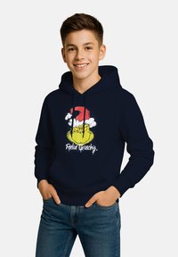 Ragazzo adolescente che sorride con le mani in tasca, indossando un felpa blu scuro con il viso del Grinch e la scritta "Mi sento Grinch".