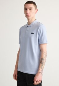 Polo bleu clair avec un col, comportant des lettres blanches, des manches courtes et un patch de marque noir sur la poitrine.