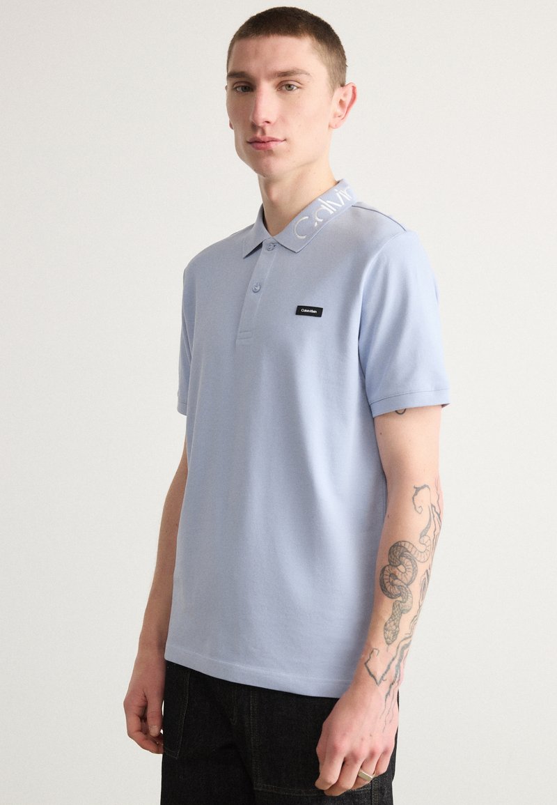 Polo bleu clair avec un col, comportant des lettres blanches, des manches courtes et un patch de marque noir sur la poitrine.