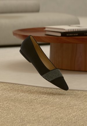 Čierna špicatá balerína s pásikom malých strieborných nitov, položená na béžovom textúrovanom koberci pred dreveným okrúhlym stolíkom a sivou pohovkou.