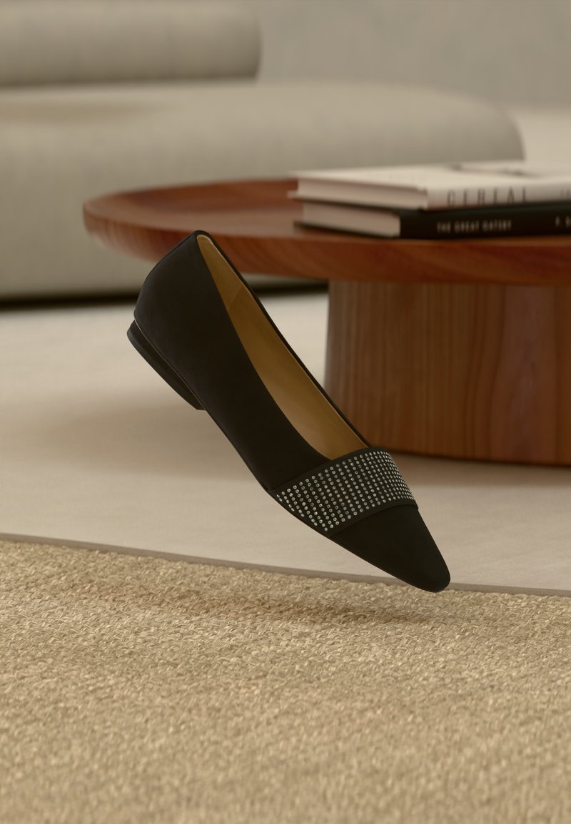 Chaussure plate noire à bout pointu avec une bande de petits clous argentés, posée sur un tapis beige texturé devant une table ronde en bois et un canapé gris.