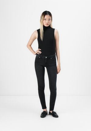 Giovane donna in piedi con una mano sui fianchi, che indossa un top senza maniche a collo alto nero, jeans skinny neri e mocassini neri su sfondo bianco.