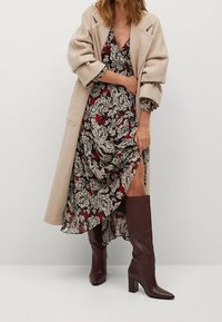 Beżowy wełniany płaszcz na wzorzystej sukience z czerwonymi i czarnymi liśćmi, połączony z brązowymi kozakami na wysokim obcasie do kolan. Sukienka ma boczne rozcięcie.