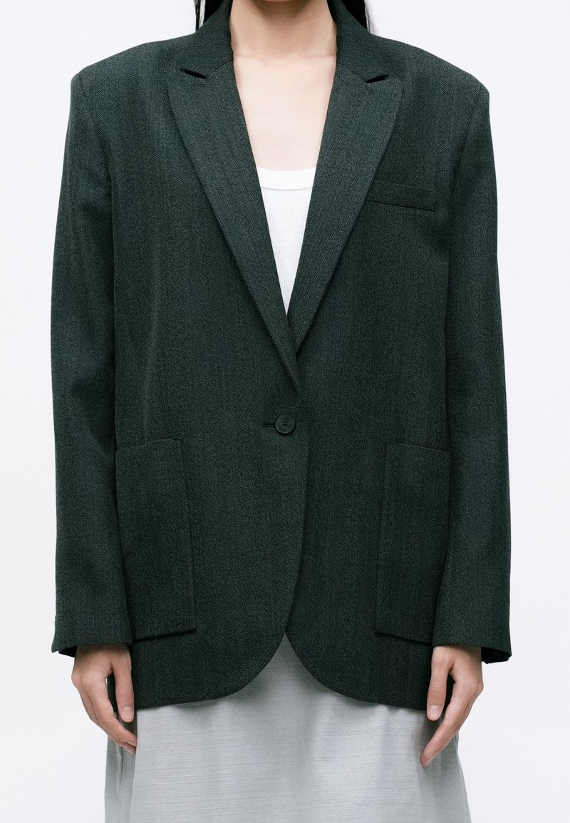 Blazer gris foncé à bouton unique avec des revers crantés et des poches plaquées, porté sur un top blanc à côtes et une jupe gris clair.