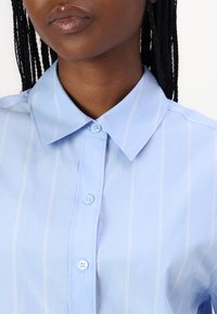 Light blue, button-down shirt featuring a classic collar and white vertical stripes. Smooth texture with two buttons visible at the collar.