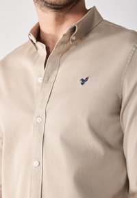 Camicetta beige con colletto classico, caratterizzata da un piccolo logo a ricamo blu di un uccellino sul petto e bottoni bianchi. Tessuto morbido.
