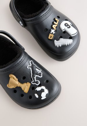 Črne Crocsi iz gume z mehko oblogo, okrašeni s športno tematskimi dodatki: pokal, nogometna žoga, igralec, dres in nogometni čevelj.