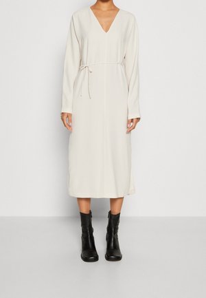 Femme portant une robe midi crème à manches longues avec col en V et fine ceinture à la taille, associée à des bottes en cuir noires mi-mollet sur fond uni.