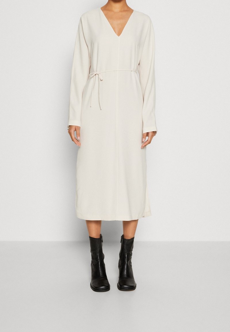 Femme portant une robe midi crème à manches longues avec col en V et fine ceinture à la taille, associée à des bottes en cuir noires mi-mollet sur fond uni.