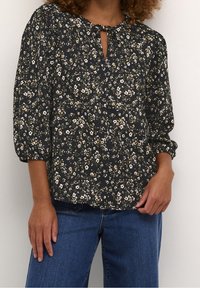 Sort bluse med blomstermønster, der har en åbning ved halsen, puffede ærmer og et struktureret stof med små hvide og gule blomster.