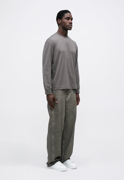 Gara piedurkne, pelēka kokvilnas krekls ar mazu logo, apvienots ar brīvām, gaiši zaļām džinsu biksēm un baltām sporta apaviem. Vienkāršs, neizstrādāts dizains.