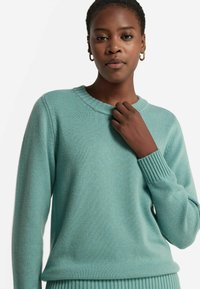 Jasnoniebieski sweter z dzianiny z prążkowanym okrągłym dekoltem i mankietami; miękka tekstura; dopasowany krój; ręka modelki delikatnie trzyma dekolt.