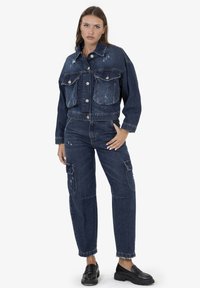 Giacca di jeans blu scuro con due grandi tasche frontali, chiusura con bottoni e dettagli consumati. Abbinata a jeans cargo coordinati e mocassini neri.