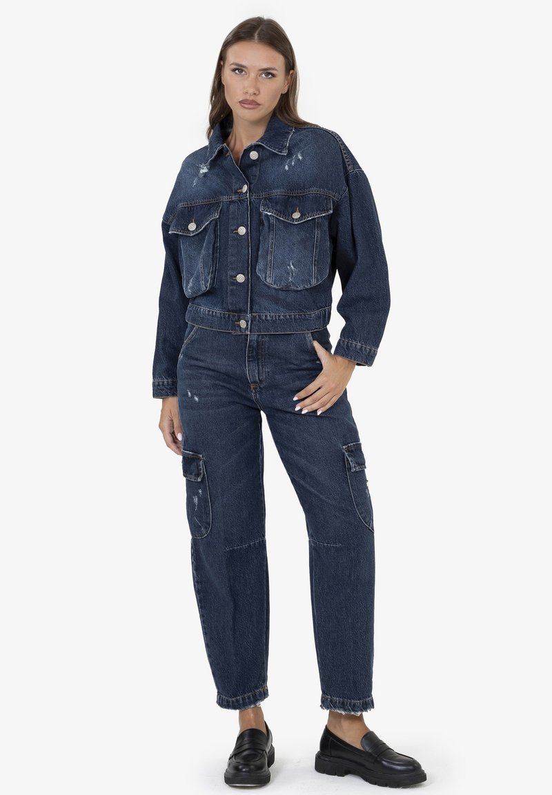 Giacca di jeans blu scuro con due grandi tasche frontali, chiusura con bottoni e dettagli consumati. Abbinata a jeans cargo coordinati e mocassini neri.