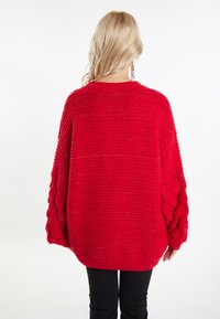 Fetzigem rotem Strickpullover mit rundem Halsausschnitt, oversized Schnitt, strukturierten Blasenärmeln und geripptem Saum. Getragen mit schwarzen Hosen.