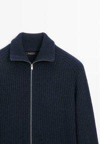 Maglione zip-up blu navy realizzato in materiale a coste testurizzato, con colletto alto e zip argentata, con un'etichetta visibile all'interno del colletto.