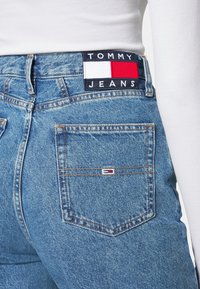 Höga blå denimjeans med en strukturerad yta, prydda med en svart och röd Tommy Jeans-logopatch vid midjan och bakfickan.