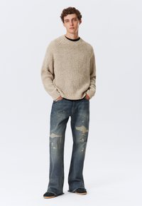 Pull en tricot beige avec des poignets et un ourlet côtelés, associé à un jean en denim bleu déchiré avec des éclaboussures de peinture et des ourlets effilochés. Chaussures noires.