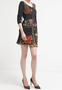 Czarna sukienka z długimi rękawami, ozdobiona kolorowym haftem kwiatowym. Sylwetka w kształcie litery A, zestawiona z małą czarną torebką na ramię i czarnymi szpilkami.