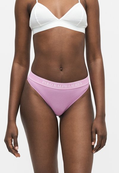 Femme portant un bralette côtelé blanc et un sous-vêtement côtelé rose avec le logo Polo Ralph Lauren sur la ceinture, debout sur fond blanc.