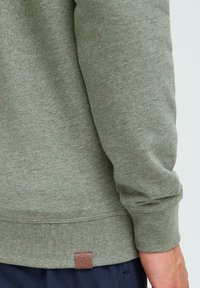 Grønn hettegenser med strukturert stoff, ribbestrikkede mansjetter og hem, med et brunt logo merkelapp på bunnen. Sidesyn av ermet.