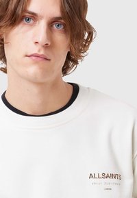 Felpa bianca con collo nero, caratterizzata dal logo "ALLSAINTS" ricamato in missione metallica sul petto. Tessuto morbido.