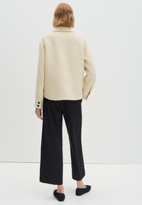 Giacca in lana beige chiaro con colletto strutturato, bottoni neri, abbinata a pantaloni neri larghi e scarpe basse nere.