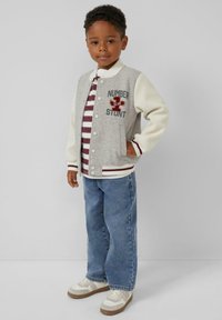 Grijze en witte varsityjack met tekst- en patchdetails, gestreept shirt, blauwe spijkerbroek en witte sneakers met beige accenten.