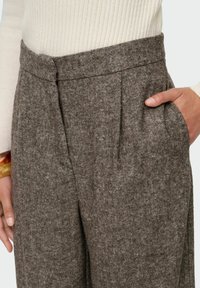 Kobieta trzyma rękę w kieszeni brązowych spodni woskowych o wysokim stanie, nosząc kremowy sweter z prążkowanej dzianiny i bransoletkę w kolorze żółwia na nadgarstku.