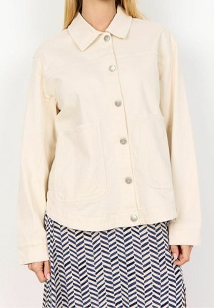 Femme portant une veste crème à boutons avec deux poches avant sur une jupe à motifs bleue et blanche.