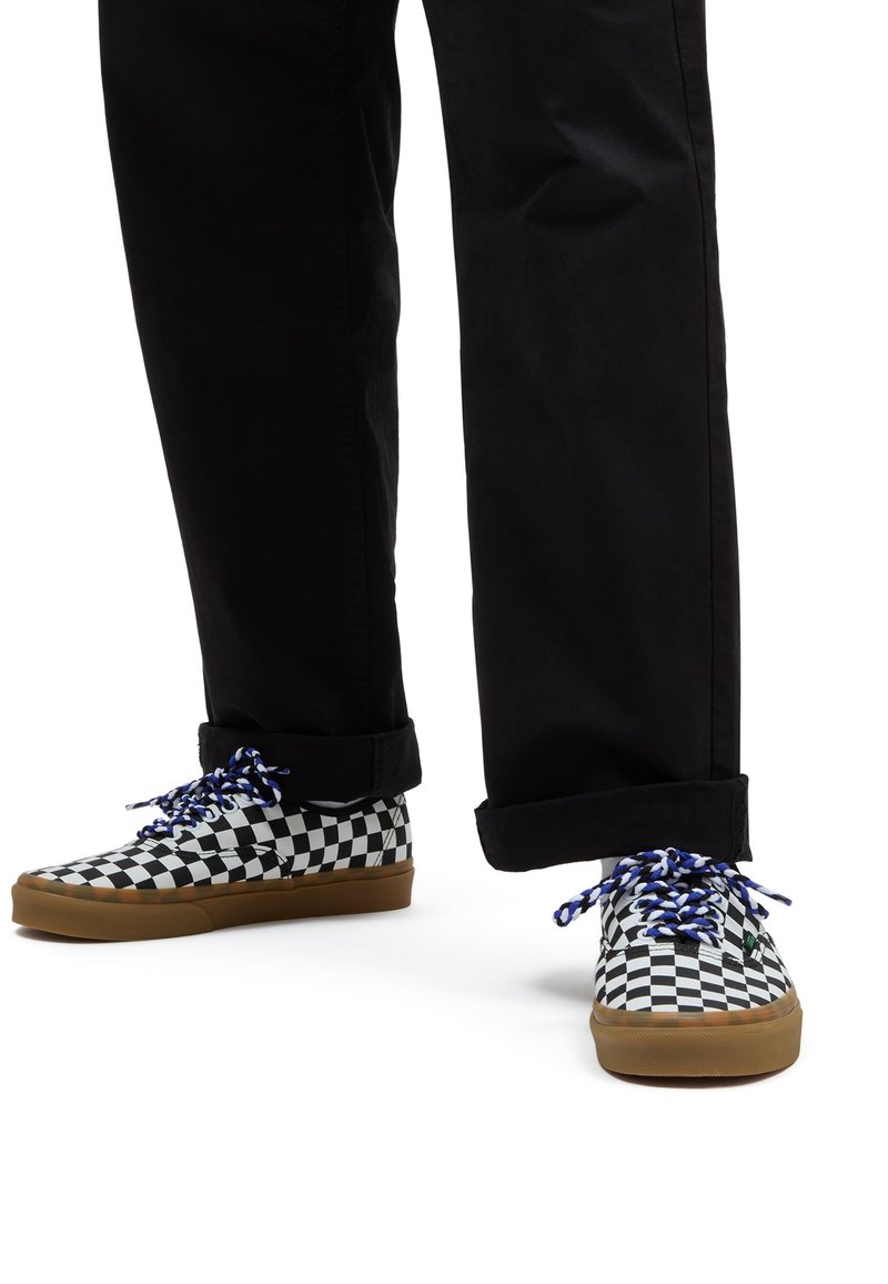 Jambes portant un pantalon noir et des baskets à damier noir et blanc avec des lacets bleus et blancs sur fond blanc.