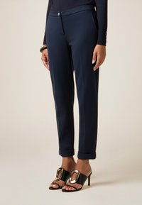 Pantaloni sartoriali blu navy slim fit, con chiusura a un bottone e orli arrotolati, abbinati a tacchi aperti neri con un accento metallico.