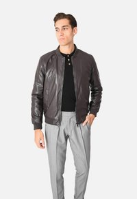 Giubbotto bomber in pelle marrone con polsini e colletto a coste, frontale con zip, abbinato a pantaloni grigi chiari plissettati. Texture liscia, design aderente.