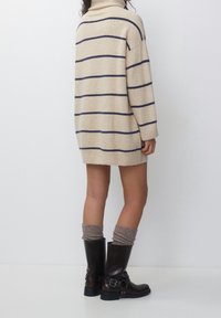 Femme portant une robe-pull à col roulé beige avec des rayures bleu marine, des chaussettes gris amples et des bottes mi-mollet marron foncé avec des boucles.