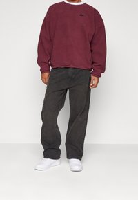 Sudadera de forro polar color burdeos con cuello redondo y logo negro, combinada con vaqueros negros de corte suelto y zapatillas blancas. Look sencillo y casual.