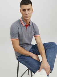 Pilkas polo marškinėliai su oranžine apykakle ir rankovių akcentais, derinami su mėlynu džinsiniu kelnių ir baltais sportbačiais. Modelis sėdi ant kėdės.