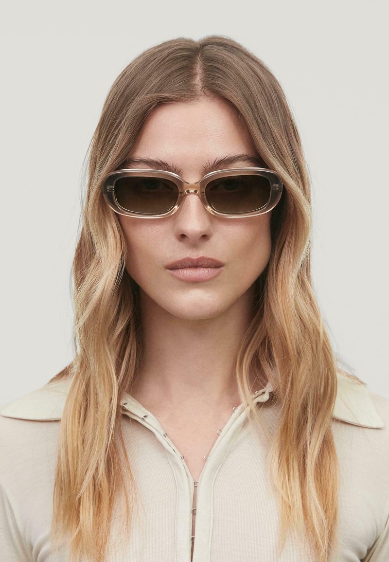 Lunettes de soleil avec des verres rectangulaires teintés de marron, monture en plastique avec des accents dorés, affichant un design moderne et une texture lisse.