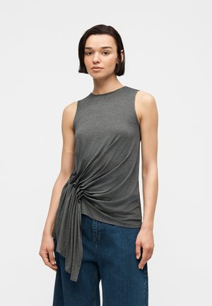 Femme aux cheveux courts et foncés portant un haut gris sans manches avec un détail froncé sur le côté et un jean bleu large, debout devant un fond uni.