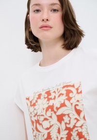 Weißes T-Shirt mit kurzen Ärmeln, das ein großes Blumenmuster in Beige und Koralle zeigt. Oben über dem Design steht der Text "Zeitlose Schönheit in voller Blüte".