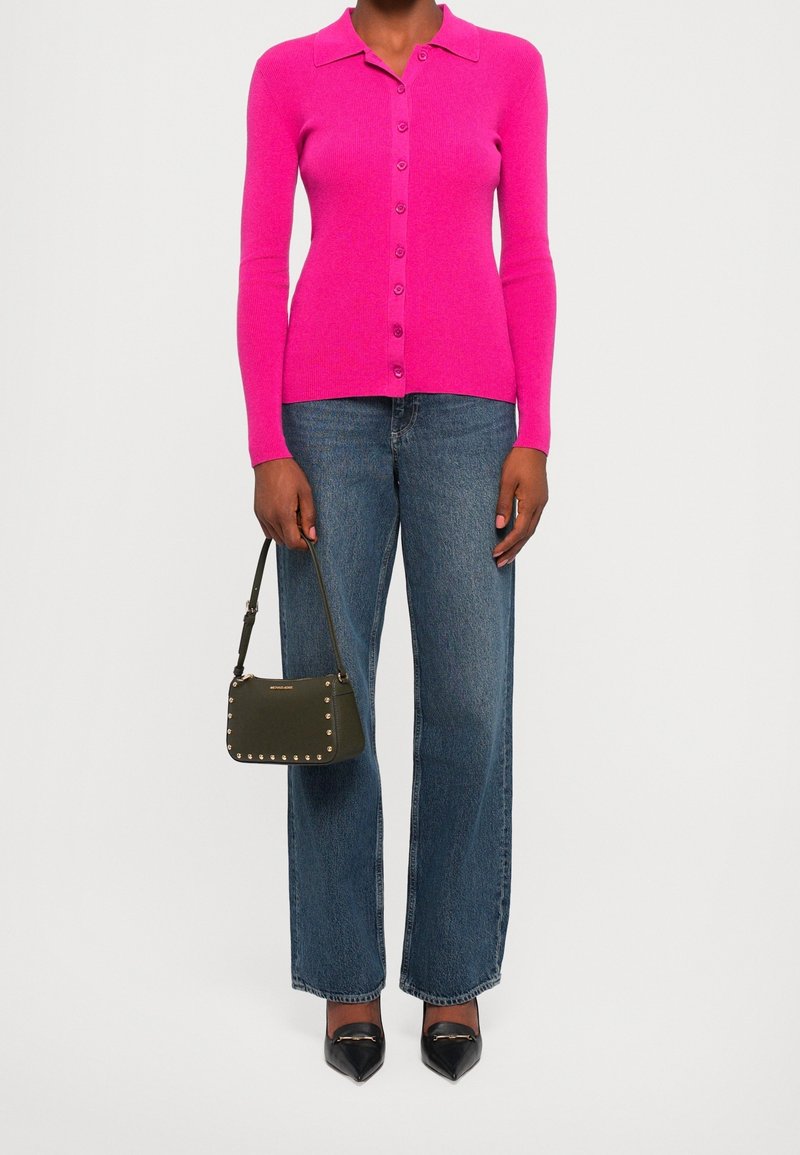 Personne portant un cardigan rose vif boutonné, un jean droit bleu, des chaussures noires pointues, et tenant un petit sac à main clouté.