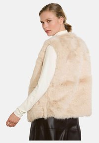 Gilet in pelliccia sintetica di un beige chiaro, con una vestibilità comoda, texture morbida e senza maniche. Indossato sopra una maglietta bianca a maniche lunghe.