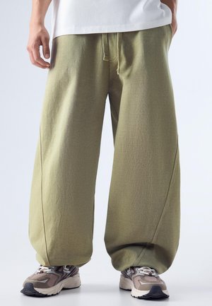 Persoană purtând pantaloni largi de culoare verde măsliniu, cu talie cu șnur, și adidași gri și bej cu model, stând pe un fundal alb.