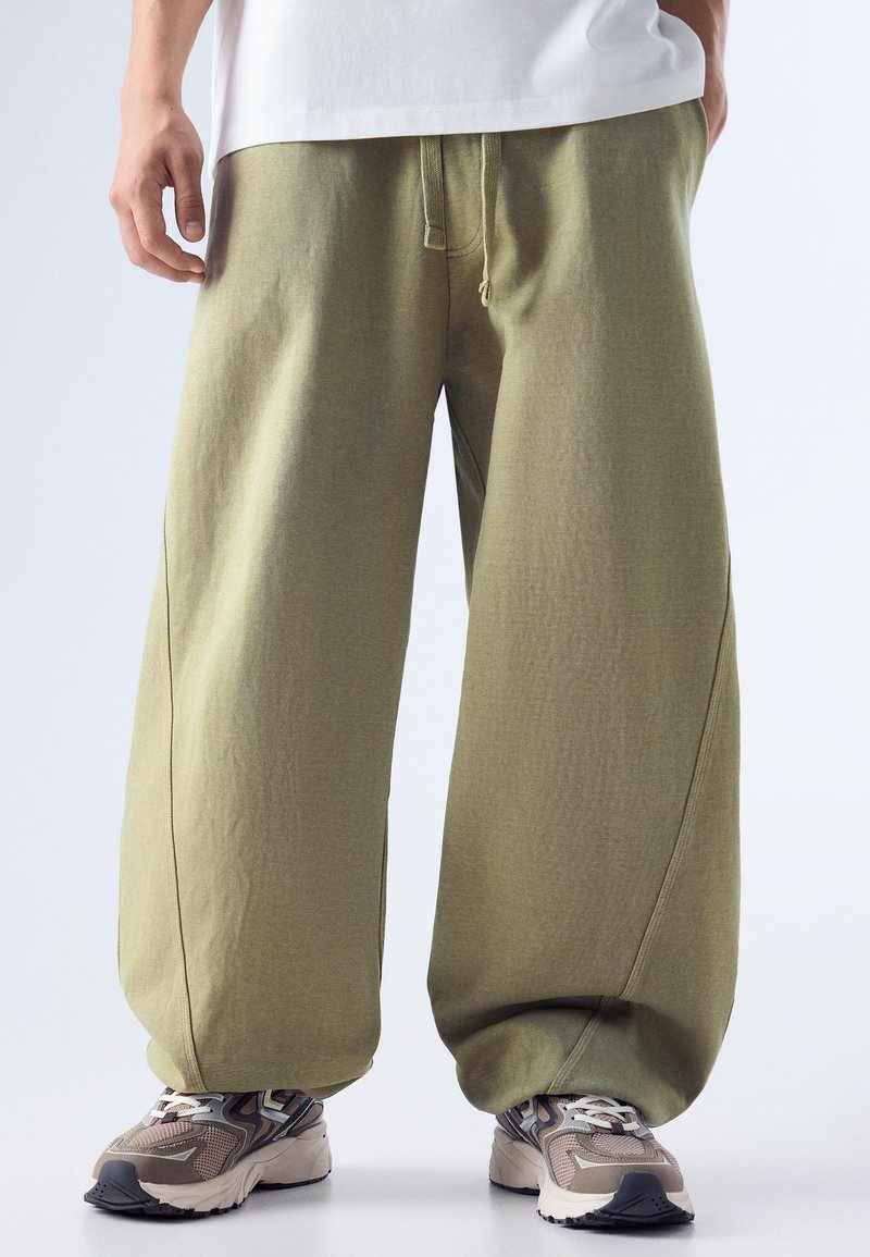 Persoon die losse olijfgroene broek draagt met een taillekoord en grijs-beige sneakers met patroon, staand op een witte achtergrond.