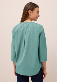 Donna con lunghi capelli castani che indossa una blusa teal larga con maniche a tre quarti e pantaloni scuri, vista di spalle su uno sfondo beige.