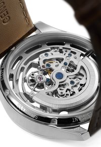 Automatisk klokke med et sølvfarget rustfritt stålkasse. Har en gjennomsiktig bakside som viser intrikate mekanismer, blå detaljer og reim i brunt lær.