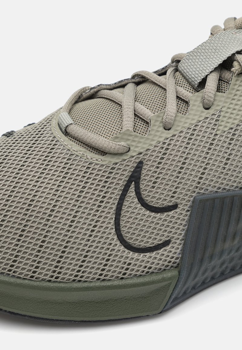 Zapatilla de malla gris claro con suela gris oscuro y acentos en verde oliva. Presenta cordones planos y un panel lateral texturizado en forma de ola.