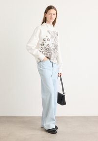Felpa bianca con un design a stampa leopardata e dettagli floreali, abbinata a jeans a gamba larga blu chiaro e scarpe nere, mentre tiene una borsa nera.